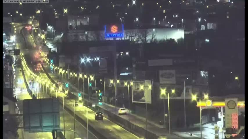 A nighttime view of a rain-slicked highway with moderate traffic, illuminated by streetlights, passing in front of buildings and a large Suzuki sign.