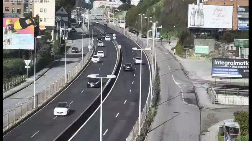 A multi-lane highway with light traffic curves gently through a city under sunny conditions, flanked by buildings and greenery.