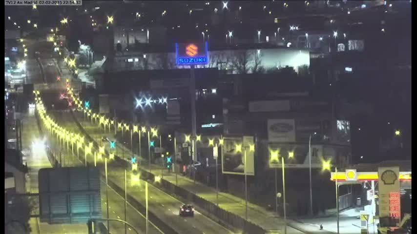 A nighttime, brightly-lit highway scene shows several cars driving, lined with streetlights and buildings on either side.