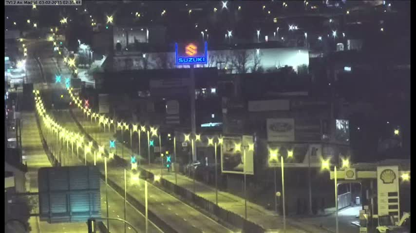 A nighttime, high-angle view of a multi-lane highway with light traffic, illuminated by streetlights, passing by buildings and a large Suzuki sign.