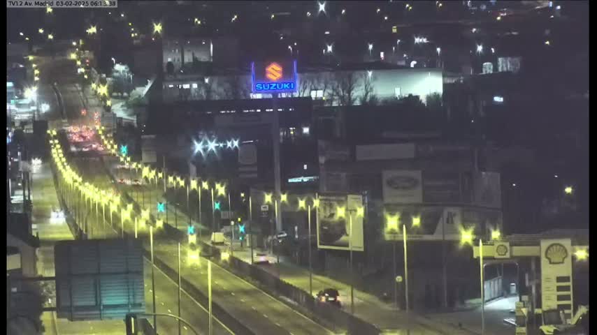 A nighttime view of a multi-lane highway with moderate traffic, lined with illuminated star-shaped lights, passing in front of various buildings and a Suzuki dealership sign.
