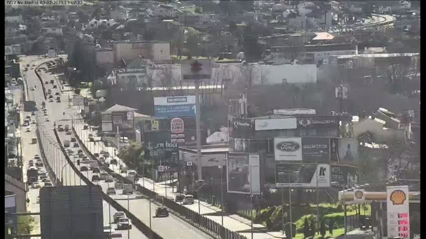 A highway with moderate traffic flows past buildings and billboards on a sunny day.