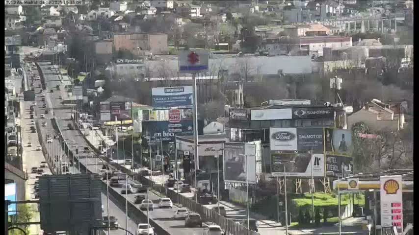 A multi-lane highway with moderate traffic flows through a city, under a bright, sunny sky.