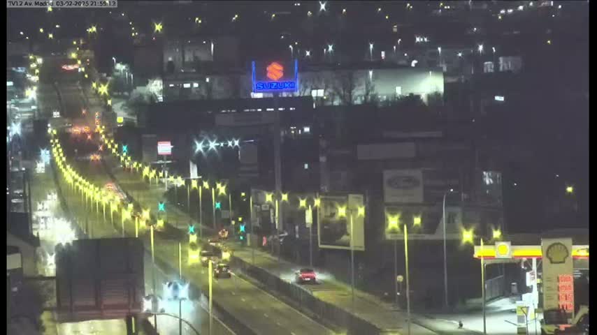 A nighttime, wet highway scene shows a multi-lane road with light traffic and illuminated streetlights, bordered by buildings and a gas station.