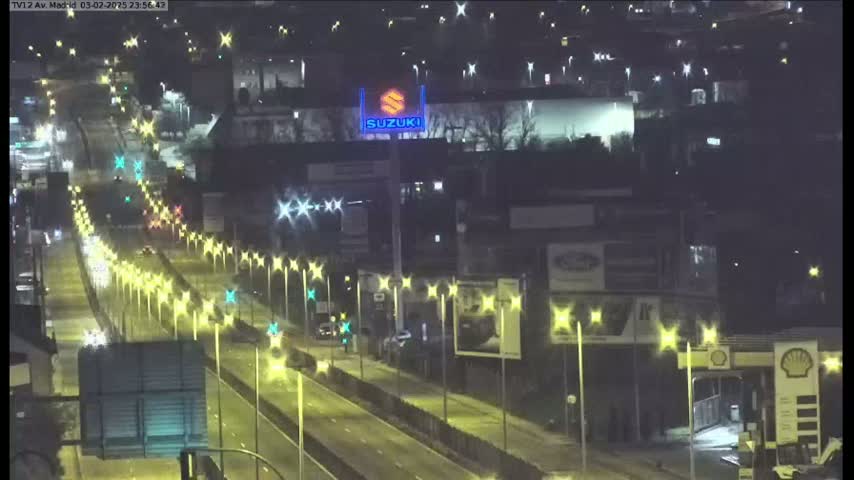 A nighttime, high-angle view shows a multi-lane highway with sparse traffic, lined with illuminated streetlights, passing by various buildings and a large illuminated Suzuki sign.