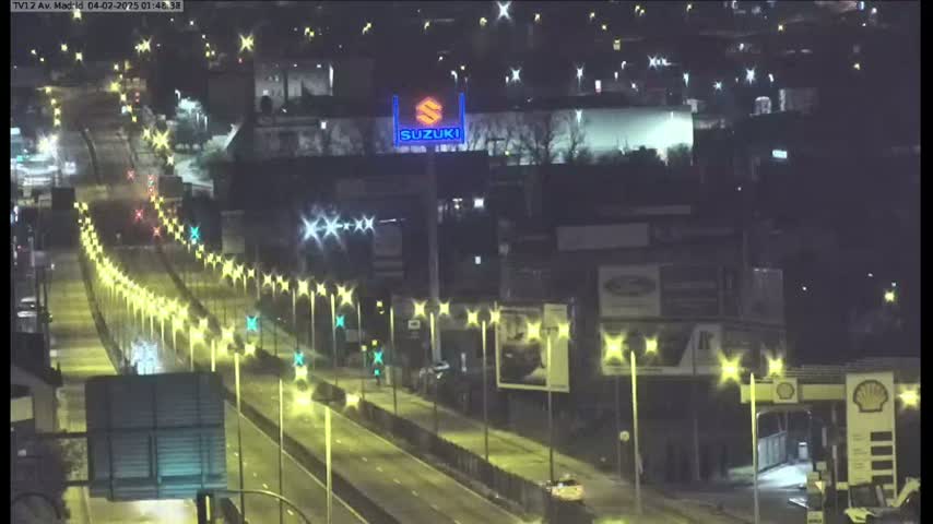 A nighttime, well-lit highway scene shows several vehicles traveling on a multi-lane road lined with streetlights, passing by buildings and a Suzuki sign.