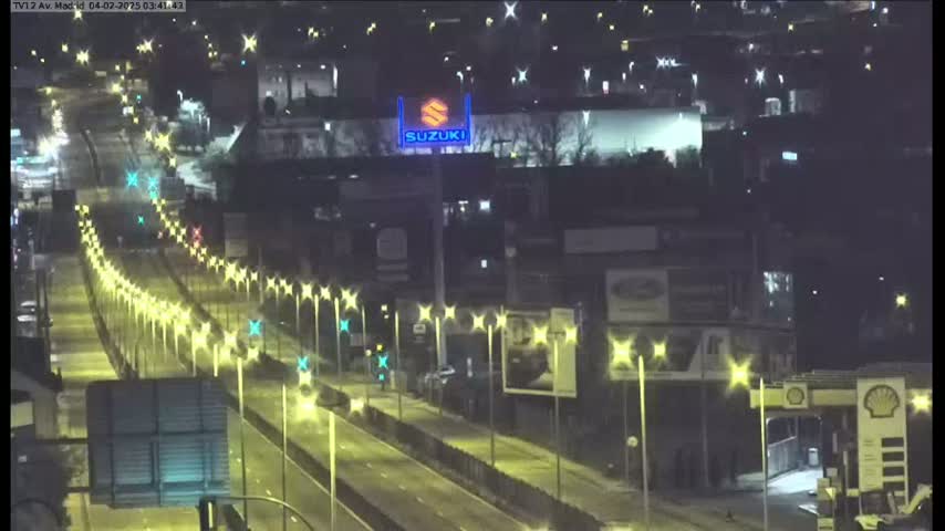 A nighttime view of a multi-lane highway with sparsely distributed traffic, lined with streetlights, and bordered by buildings and billboards.