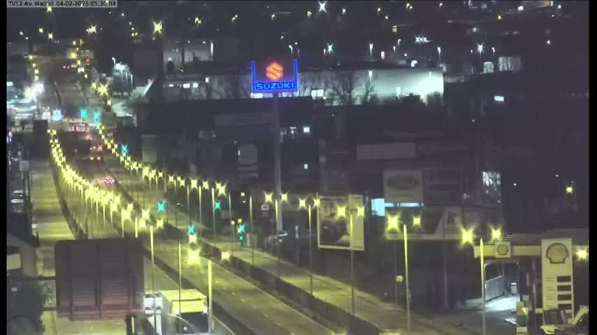 A nighttime, illuminated highway scene shows a multi-lane road with sparse traffic and streetlights lining both sides, passing in front of buildings and a gas station.
