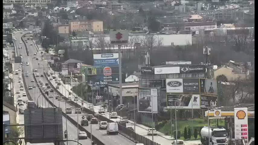 A highway scene on a sunny day shows multiple lanes of traffic moving past buildings and billboards.
