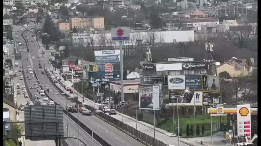 A multi-lane highway with moderate traffic flows through a city under overcast skies, lined with buildings and billboards.