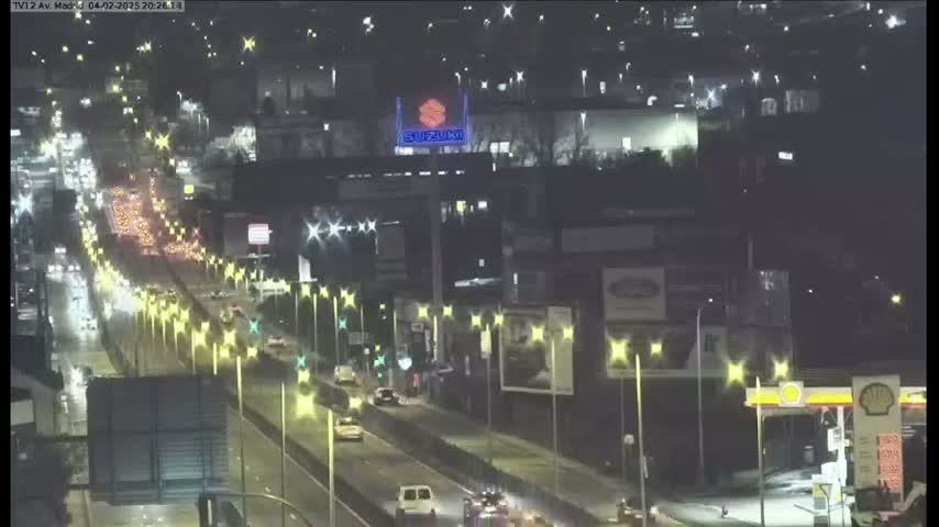 A rain-slicked highway at night is congested with numerous vehicles, surrounded by illuminated buildings and streetlights.