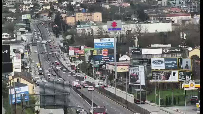 A highway with heavy traffic flows through a city under an overcast sky, lined with buildings displaying numerous billboards.