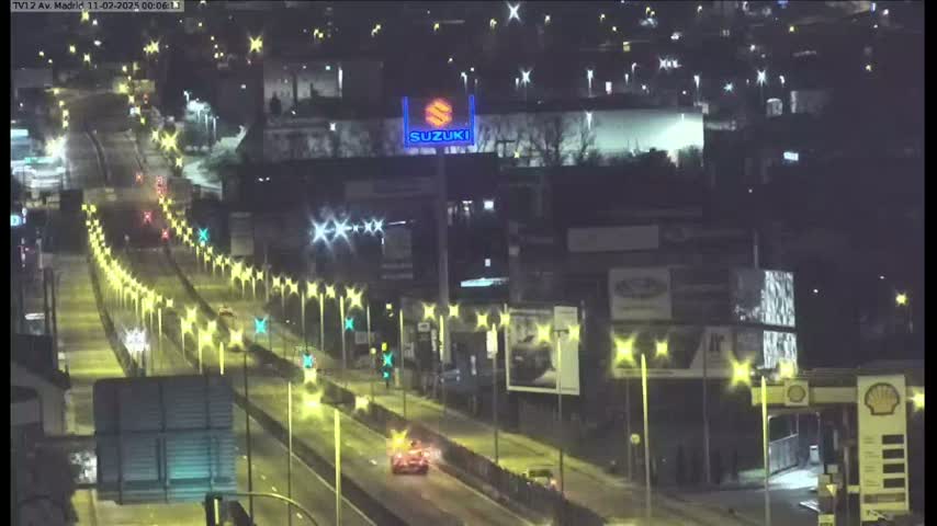 A nighttime view of a multi-lane highway with sparse traffic, lined with streetlights and flanked by buildings, including a large Suzuki sign.