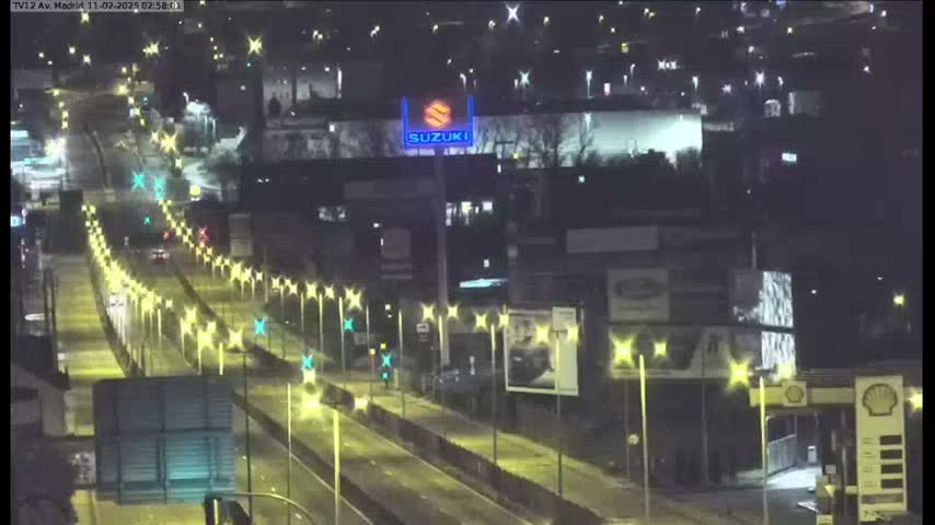 A nighttime, illuminated highway scene shows a multi-lane road with sparse traffic, lined with light posts, and buildings along the sides.