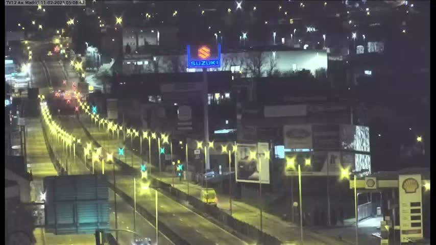 A nighttime, illuminated highway scene shows several vehicles driving on a multi-lane road lined with streetlights, passing by buildings and billboards.
