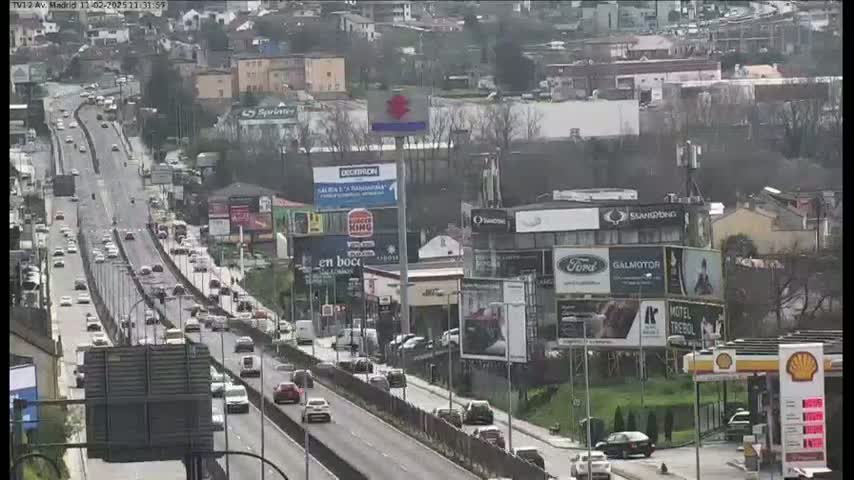 A highway scene on an overcast day shows numerous vehicles driving on a multi-lane road, passing by buildings with billboards and gas stations.