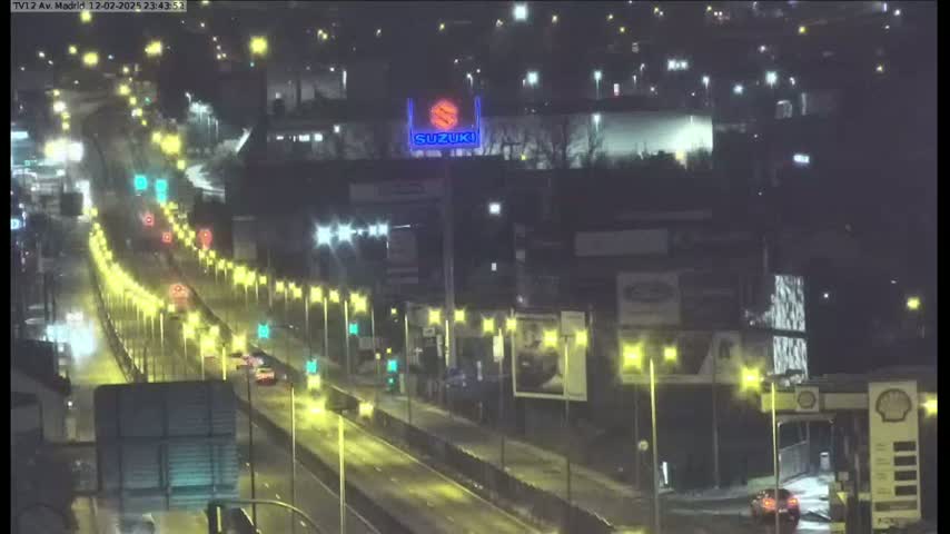 A nighttime, lightly trafficked highway scene shows several vehicles traveling under illuminated streetlights, passing by various buildings and a large Suzuki advertisement.