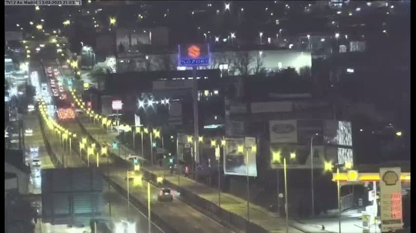 A wet, nighttime highway scene shows numerous vehicles driving on a multi-lane road lined with lights, passing buildings with illuminated signage.