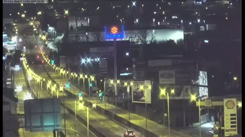 A nighttime, brightly lit highway scene shows multiple lanes of traffic moving along a multi-lane road flanked by buildings and illuminated by streetlights.