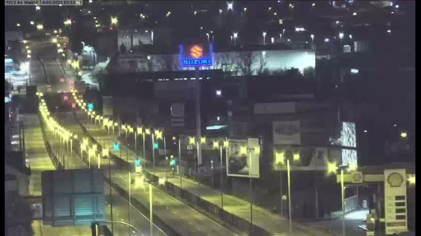 A nighttime, wet highway scene shows a multi-lane road with sparse traffic, flanked by illuminated streetlights and buildings, one of which displays a Suzuki logo.
