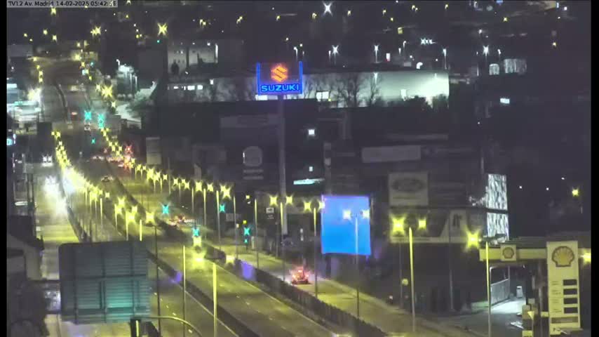 A nighttime, brightly lit highway with sparse traffic passes in front of buildings and a large Suzuki sign.