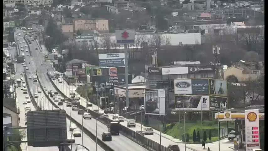 A highway scene on a partly cloudy day shows numerous vehicles traveling along a multi-lane roadway, surrounded by buildings and billboards.
