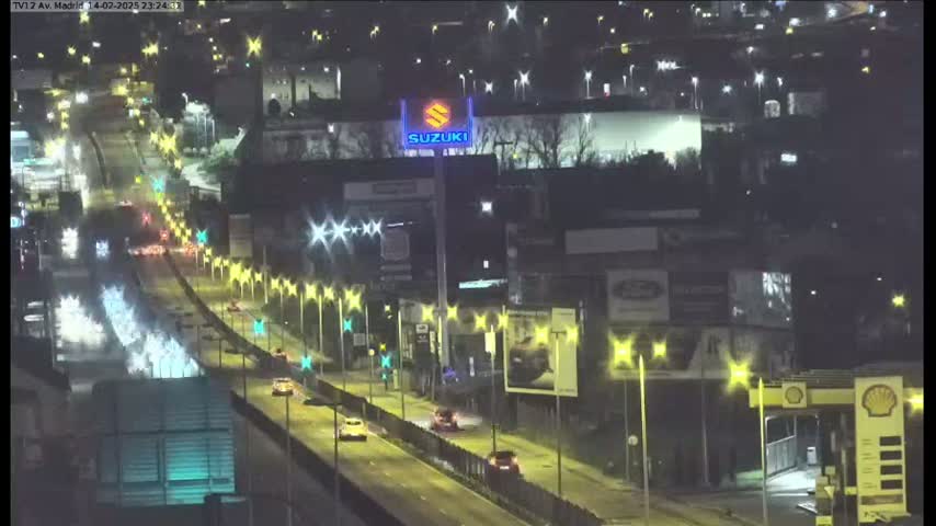 A nighttime, brightly lit highway scene shows several vehicles driving along a multi-lane road, flanked by buildings with billboards and streetlights.