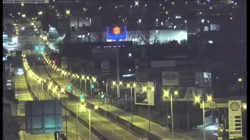 A nighttime, high-angle view of a multi-lane highway with sparse traffic, lined with illuminated streetlights and flanked by buildings and billboards.