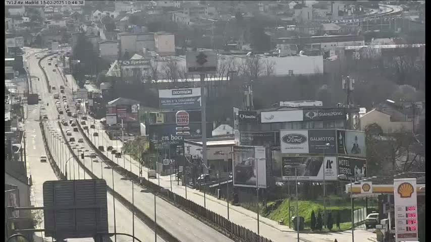A highway with moderate traffic flows through a city on a sunny day, lined with buildings and billboards.