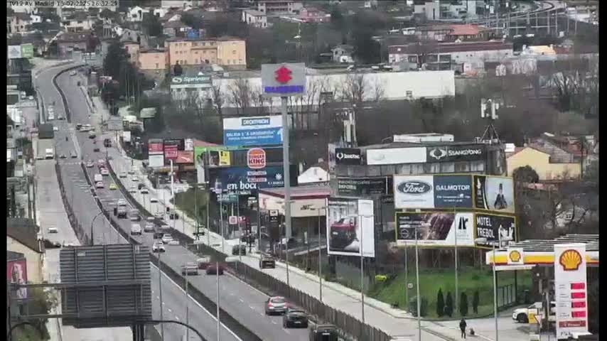 A highway with moderate traffic flows through a city on a partly cloudy day, passing numerous billboards and businesses.