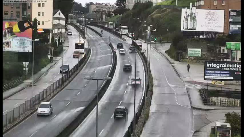 A wet, multi-lane highway curves gently, carrying several cars and a bus, with buildings and billboards visible along the roadside.