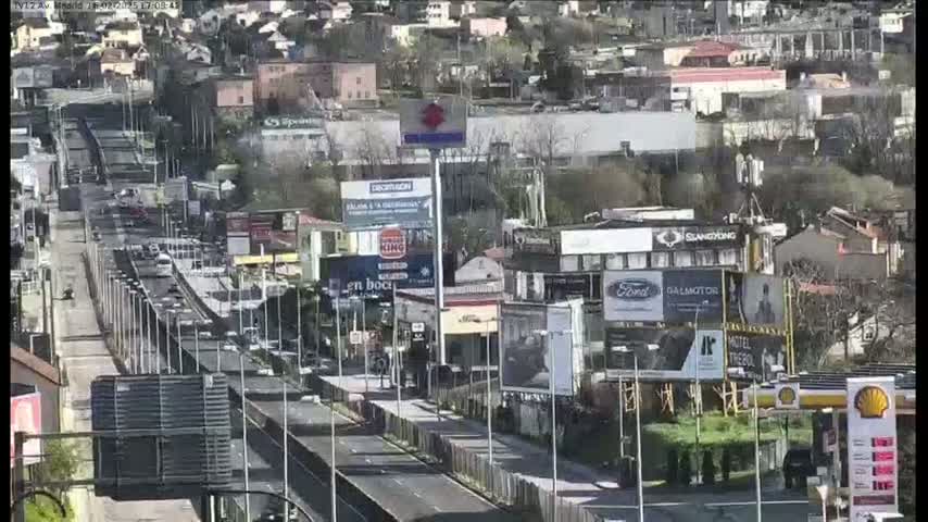 A highway with moderate traffic flows through a city on a sunny day, lined with buildings and billboards.