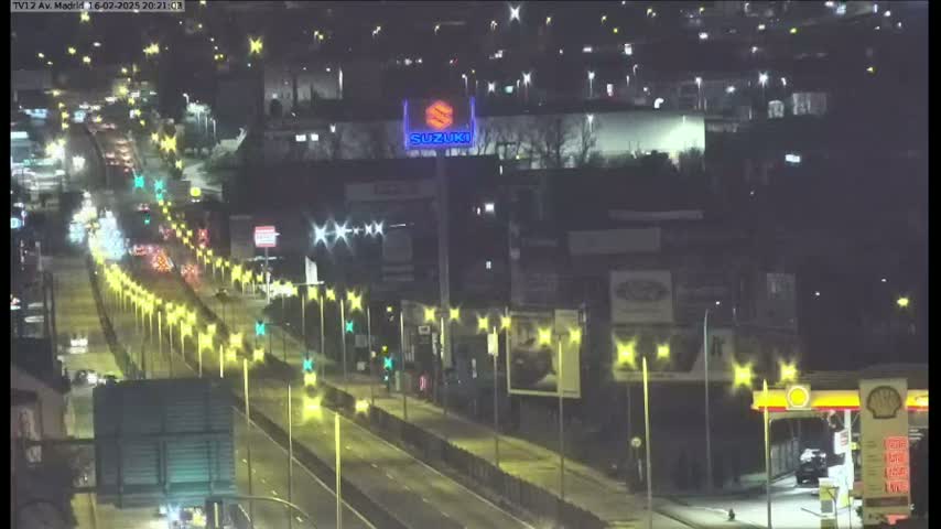 A nighttime view of a highway with moderate traffic, illuminated by streetlights, alongside buildings and a gas station.