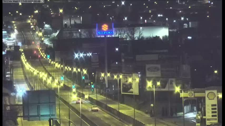 A nighttime view of a multi-lane highway with sparse traffic, lined with illuminated streetlights, passing in front of buildings and billboards.