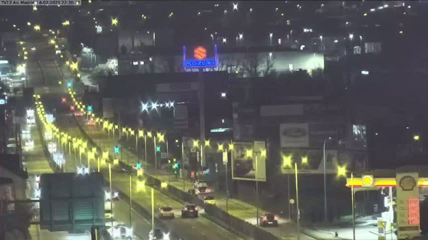 A wet, nighttime highway scene shows several vehicles traveling along a multi-lane road lit by streetlights, with buildings and advertisements visible on either side.