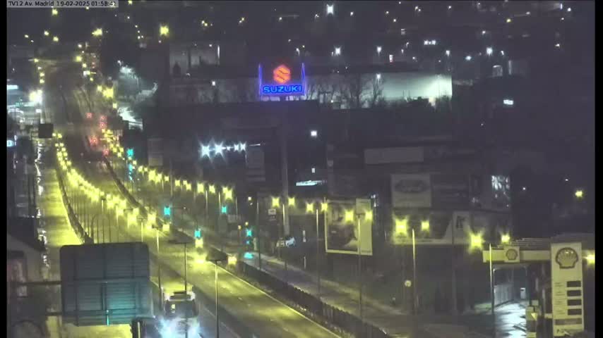 A rain-slicked highway at night is brightly lit by streetlights and shows sparse traffic passing under a Suzuki billboard.