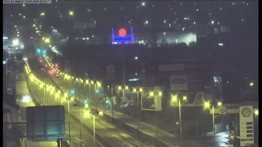 A nighttime view of a highway with moderate traffic, illuminated by streetlights, passing by buildings and a gas station under overcast skies.