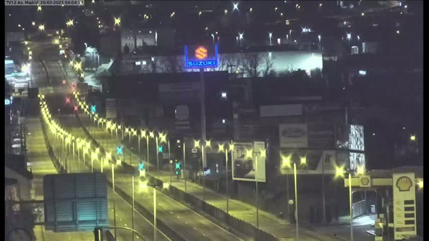 A nighttime, brightly lit highway with sparse traffic passes in front of buildings and billboards.