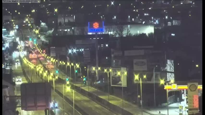 A wet, nighttime highway scene shows moderate traffic moving along a multi-lane roadway, lined with illuminated streetlights and bordered by buildings and businesses.