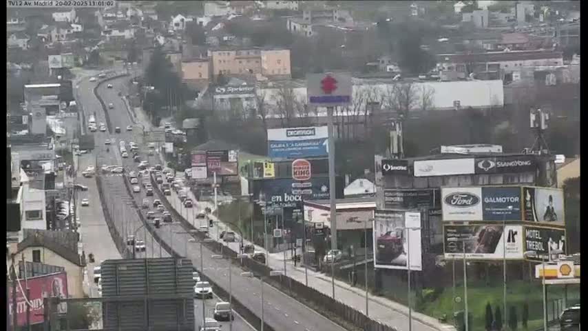 A highway with moderate traffic flows through a city on an overcast day, passing numerous billboards and buildings.