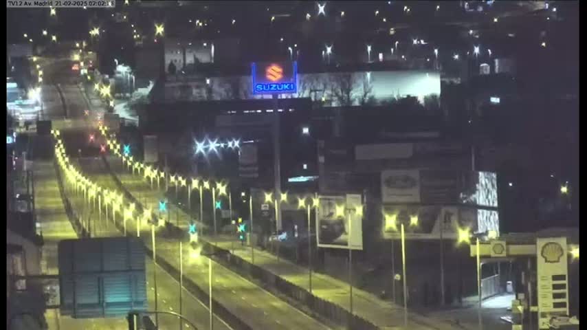 A nighttime, brightly-lit highway scene shows sparsely traveled lanes, lined with streetlights, leading towards a building displaying a Suzuki logo.