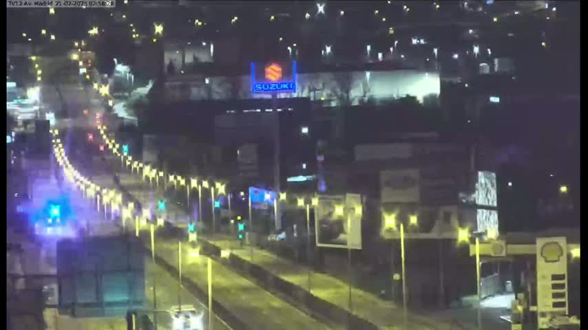A nighttime, wet highway scene shows several vehicles traveling along a multi-lane road lined with illuminated streetlights, with a large illuminated Suzuki sign visible in the background.