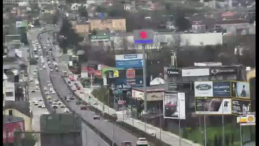 A multi-lane highway with moderate traffic flows past numerous buildings and billboards on an overcast day.