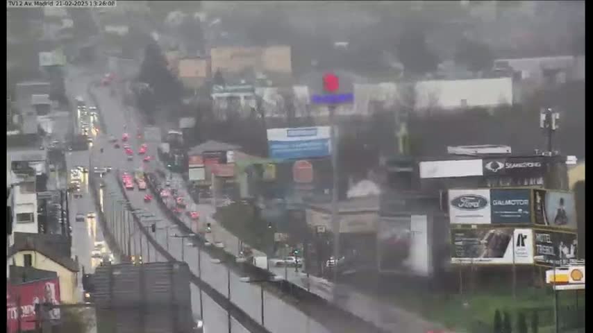 A rain-soaked highway with moderate traffic is viewed from an elevated perspective, showing buildings and billboards lining the road.