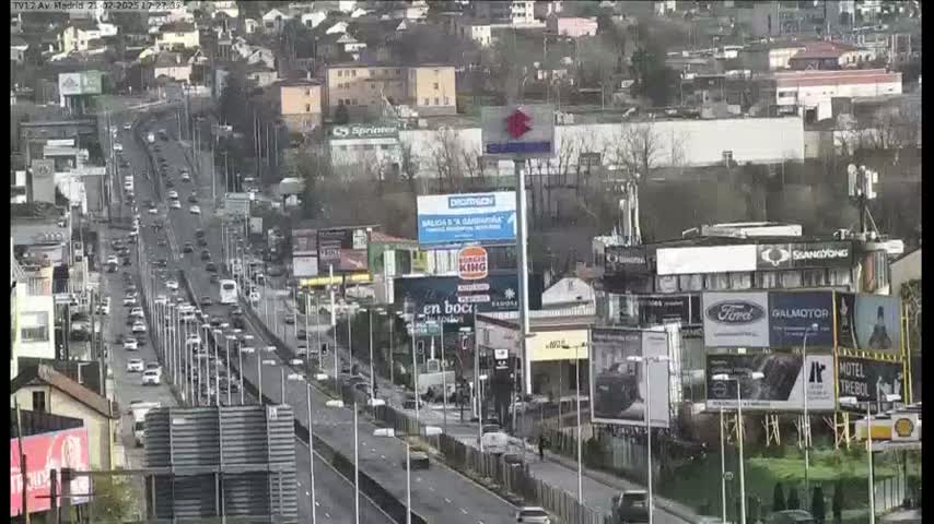 A highway with moderate traffic flows through a city on an overcast day, passing numerous billboards and buildings.