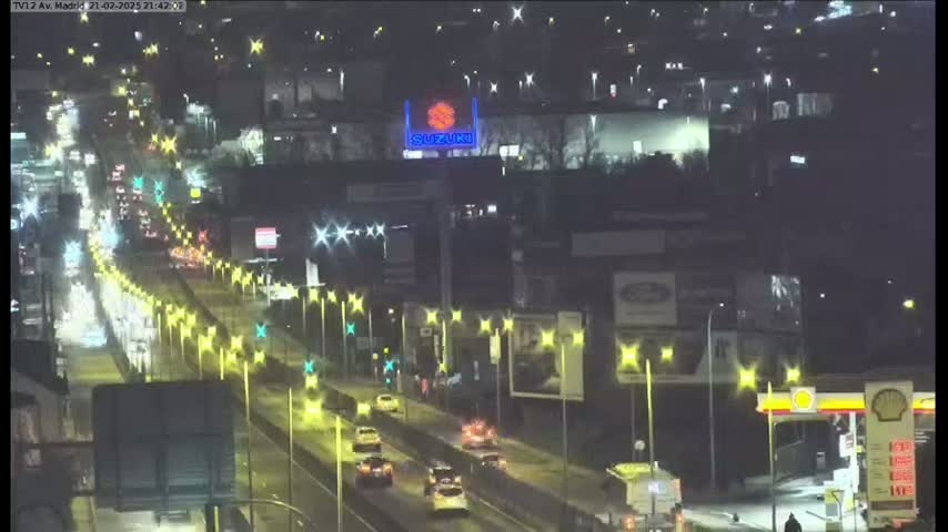 A nighttime, slightly wet highway scene shows moderate traffic moving along a multi-lane road lined with illuminated streetlights and billboards.