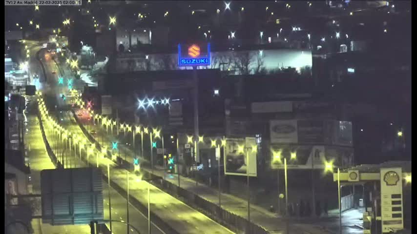 A nighttime, brightly lit highway with sparse traffic stretches into the distance, passing by buildings and a gas station.