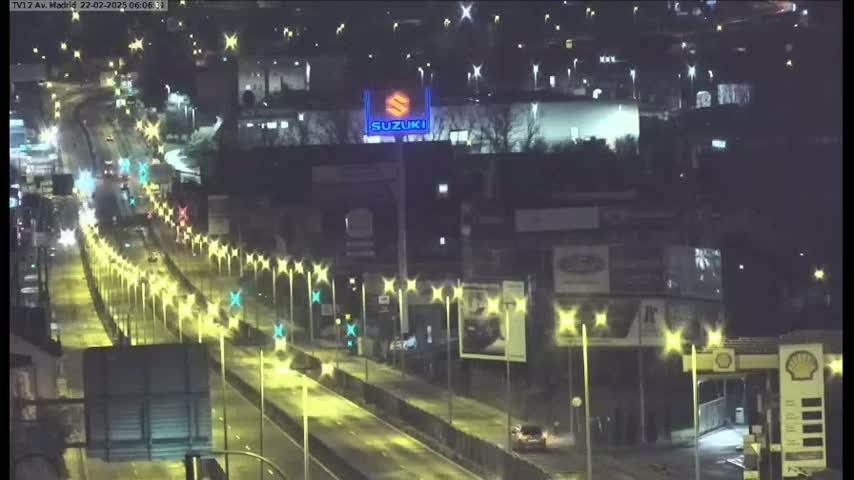 A nighttime, illuminated highway scene shows a multi-lane road with sparse traffic, lined with streetlights, passing in front of buildings and businesses.