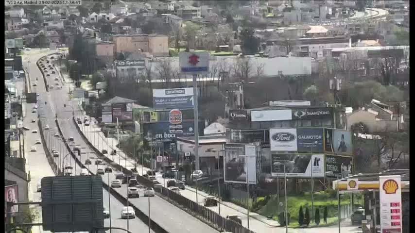 A highway scene on a partly cloudy day shows multiple lanes of traffic moving along a multi-lane roadway lined with buildings and billboards.