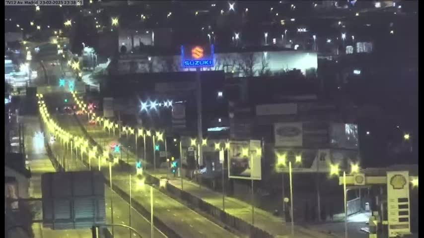 A rain-slicked highway at night is brightly lit by streetlights, with sparse traffic and buildings visible in the background.